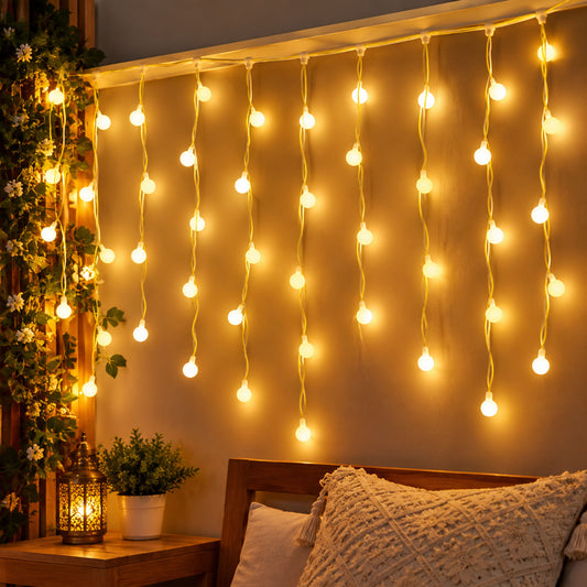 Warm white bulb string lights hang vertically on a wall above a bed with a decorative pillow and a small plant.