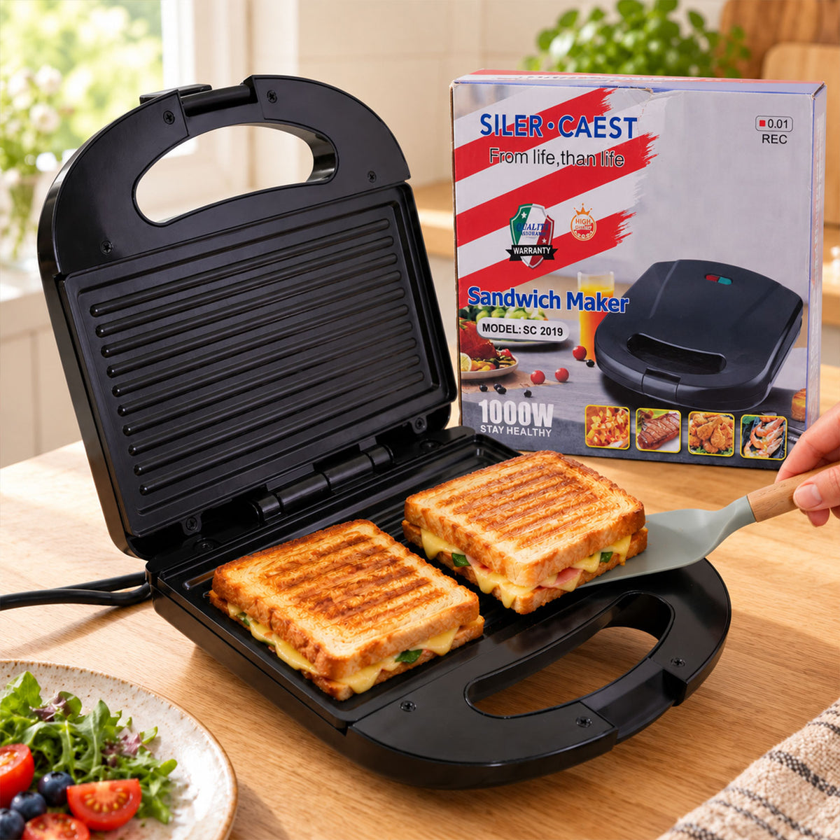 An open black sandwich maker with two grilled sandwiches inside, a spatula removing one, and its box in the background.
