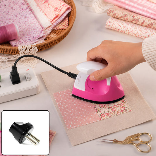 A hand holds a pink and white mini iron over a piece of floral fabric on a beige cloth, with a power strip and craft