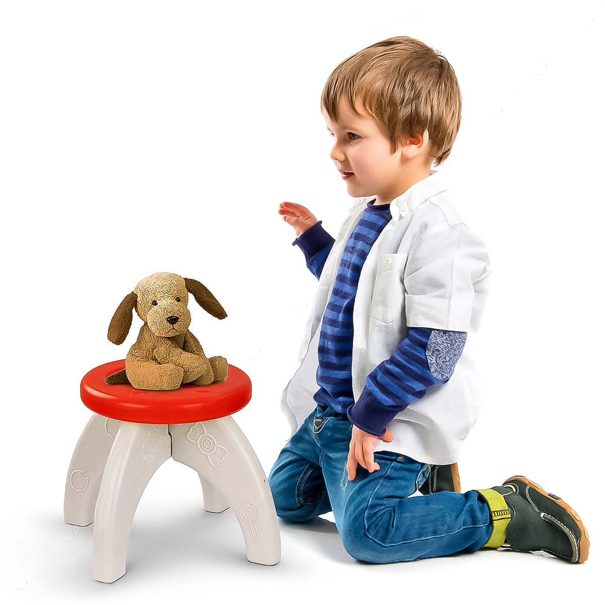 A young boy kneels beside a small red and white plastic stool with a brown stuffed dog sitting on it.