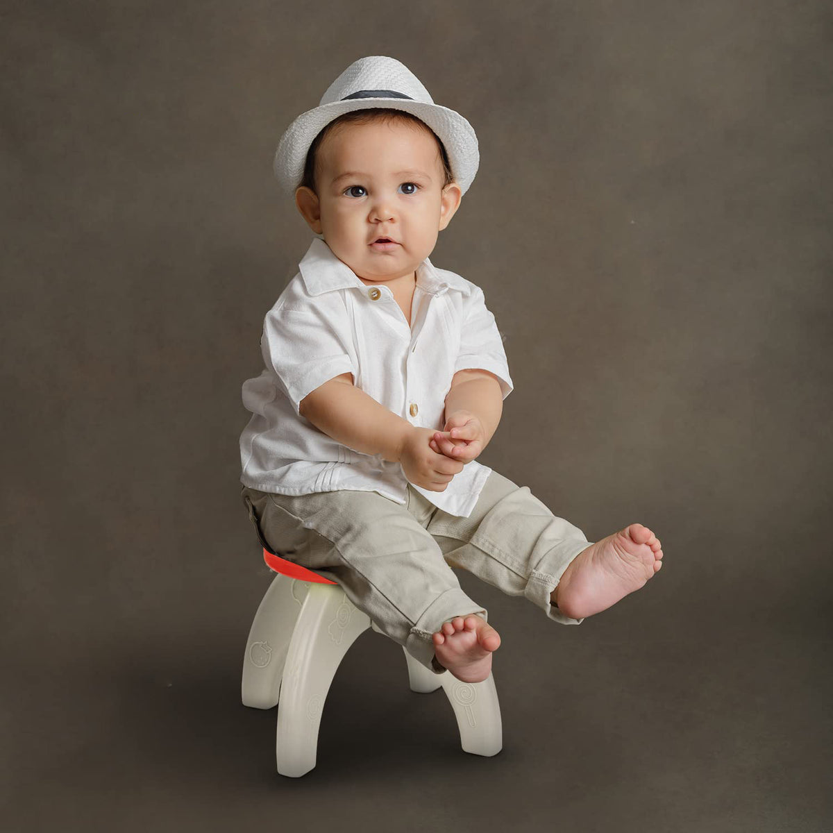 A baby wearing a white shirt and hat sits on a small red and white plastic stool against a brown backdrop.