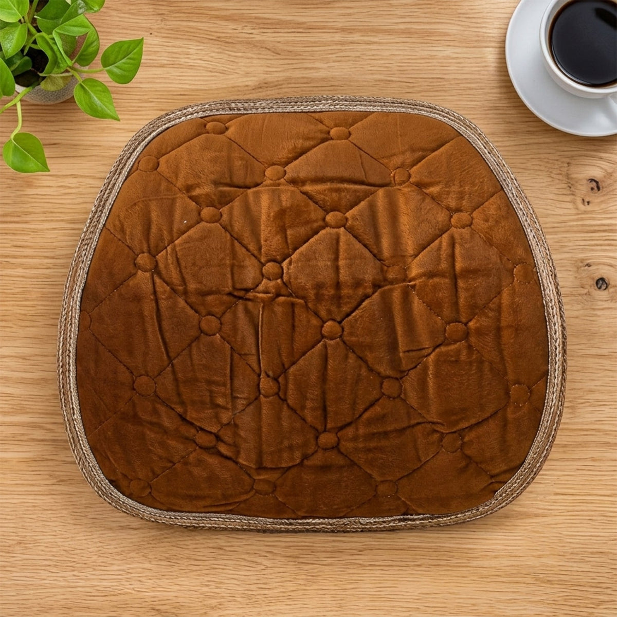Overhead view of a brown quilted velvet cushion on a wooden desk with a plant and coffee.
