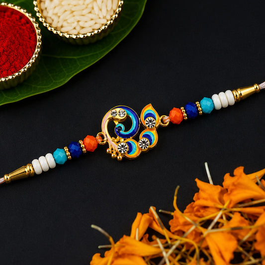 A gold and blue peacock-shaped Rakhi with orange, blue, and white beads sits on a black surface next to red powder and white