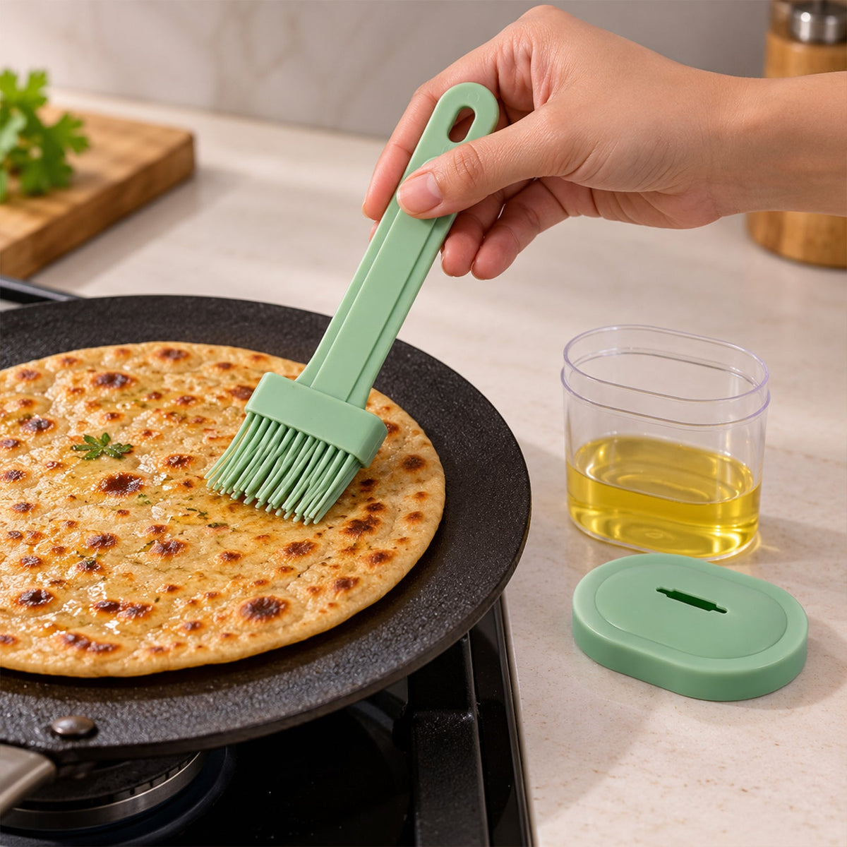 A hand uses a light green silicone basting brush to spread oil on a flatbread cooking on a black griddle, with a clear oil