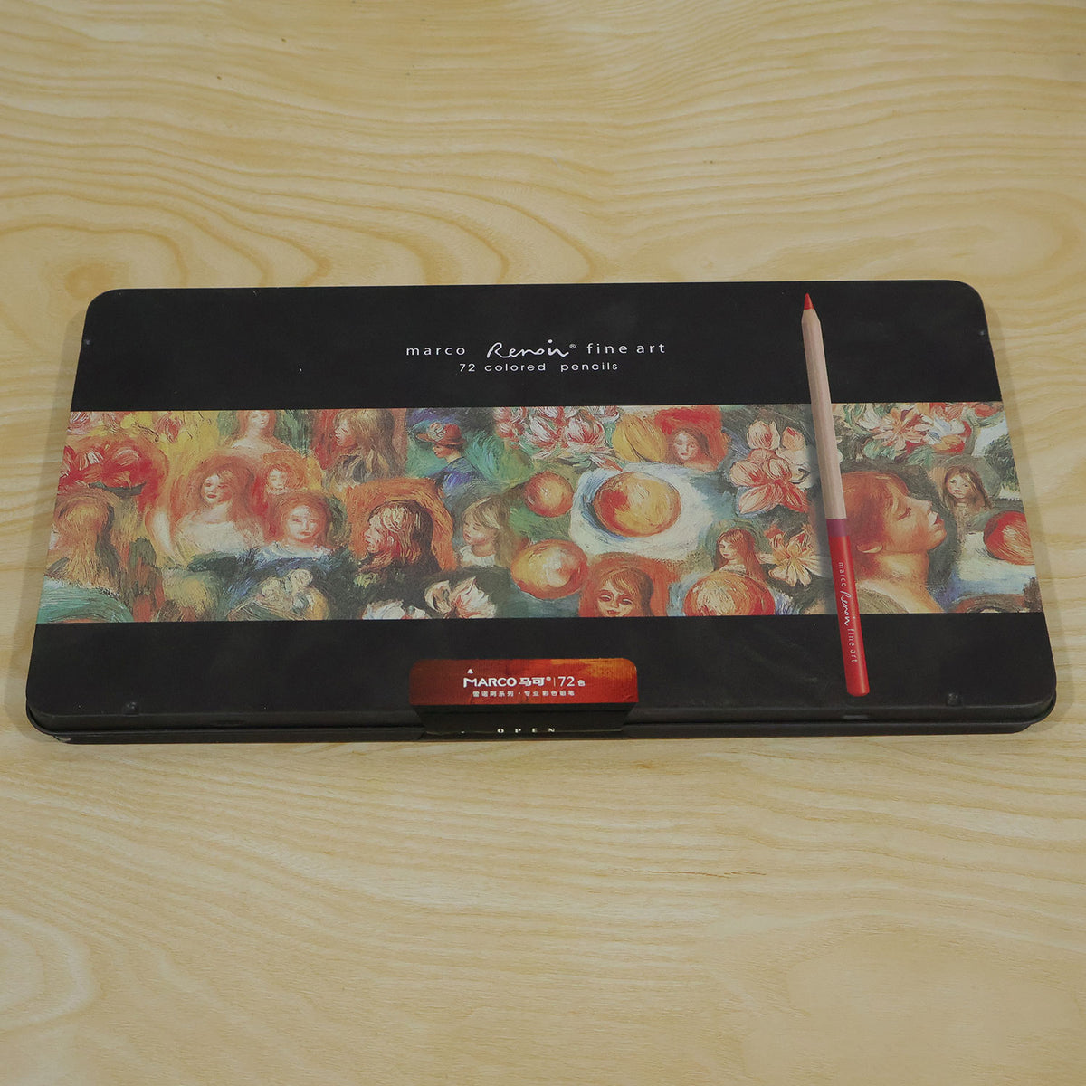 A closed black metal tin with a red colored pencil resting on top, displayed on a light wooden surface.