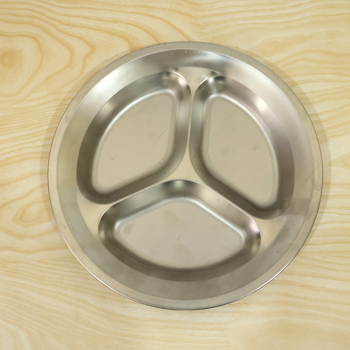 A top-down view of an empty, round, three-compartment stainless steel plate on a light wooden surface.