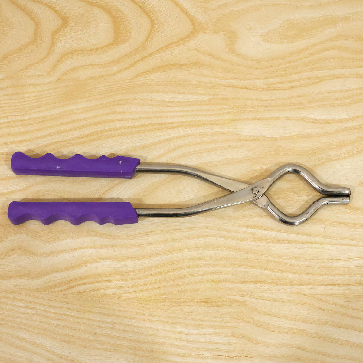 A stainless steel pot lifter with purple handles lying on a light wooden surface.