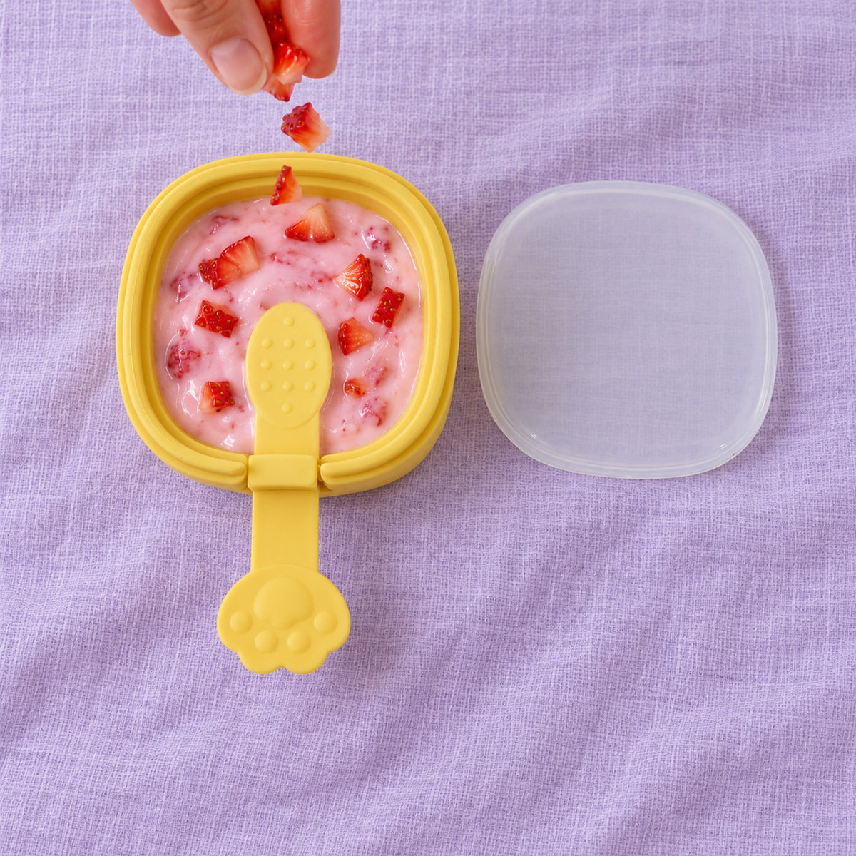 A hand drops diced strawberries into a yellow square mold filled with pink ice cream mixture, beside a clear lid.
