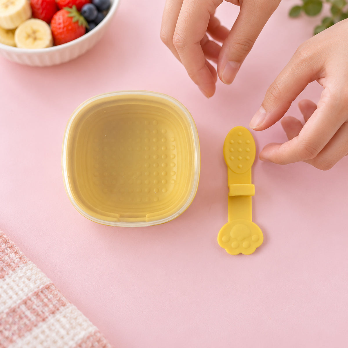 Two hands adjust a yellow paw-shaped popsicle stick next to an empty yellow square mold with a clear lid on a pink surface.
