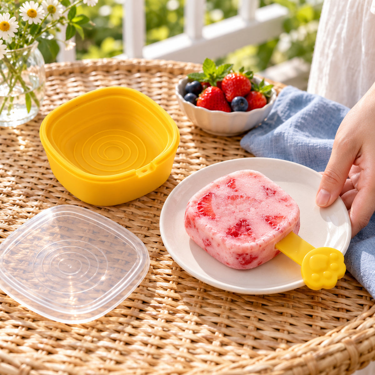 A person holding a white plate with a strawberry popsicle, next to an empty yellow mold and a clear lid.