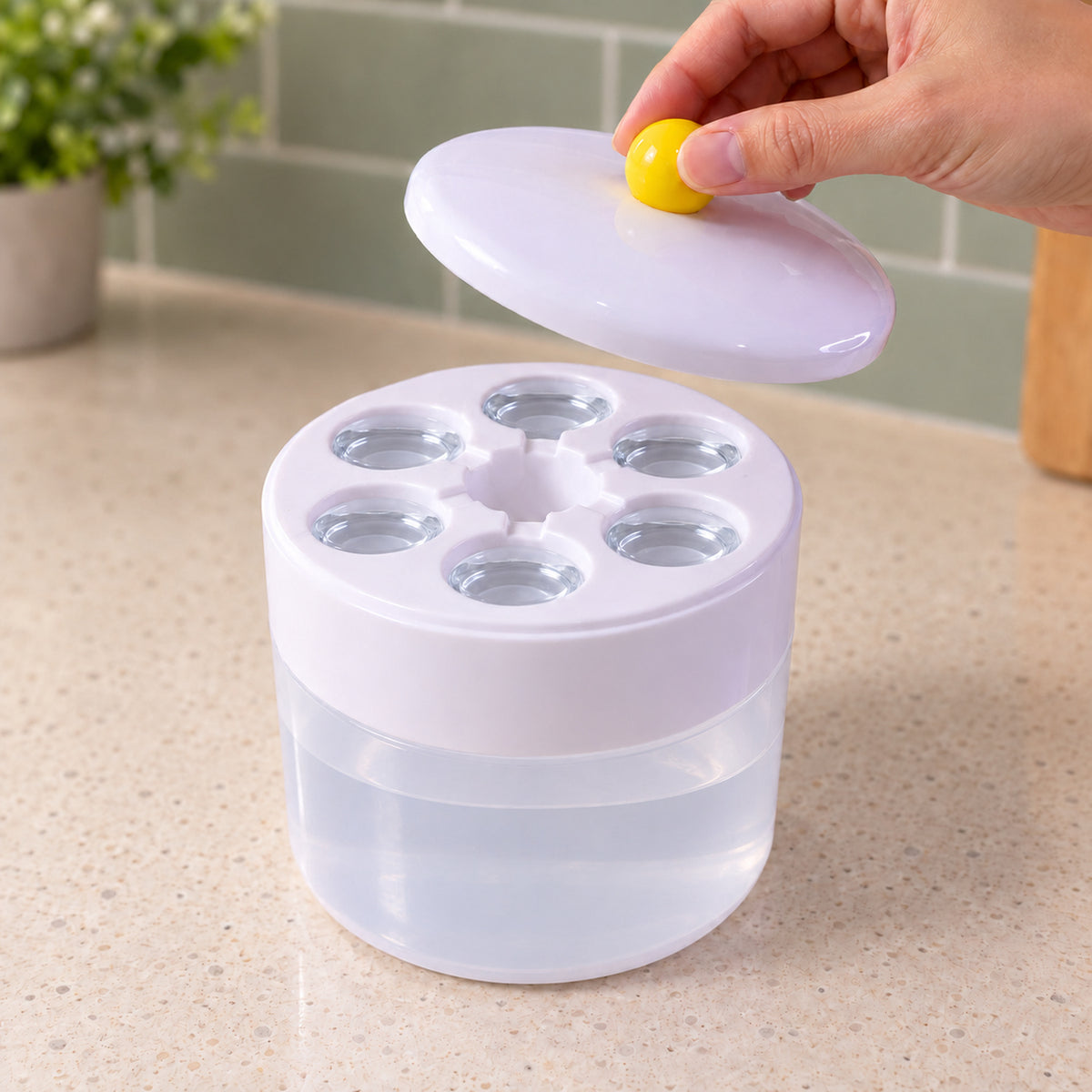 A hand lifts the white lid with a yellow knob from the ice cube maker, revealing six circular cavities filled with water.