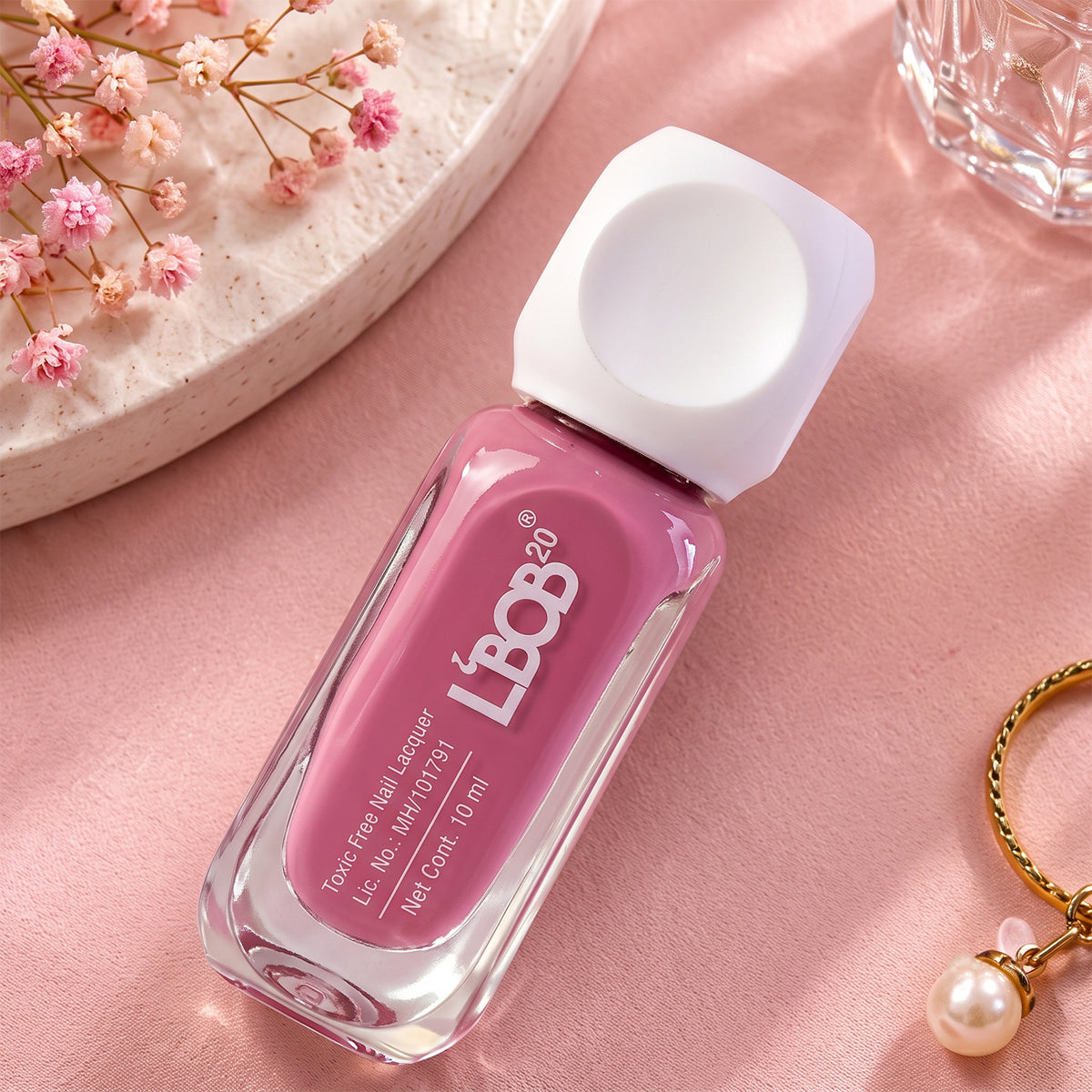 A mauve nail polish bottle rests on a pink surface next to a pearl charm and small flowers.
