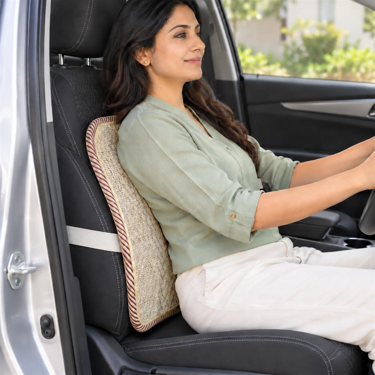 A woman with dark hair wearing a light green shirt and white pants sits in a car, using a beige mesh back support cushion.
