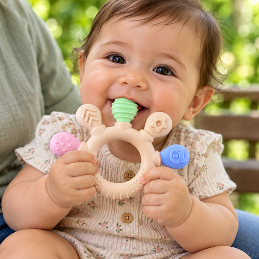 A happy baby in a floral onesie chews on a beige teether with green, pink, and blue textured ends.
