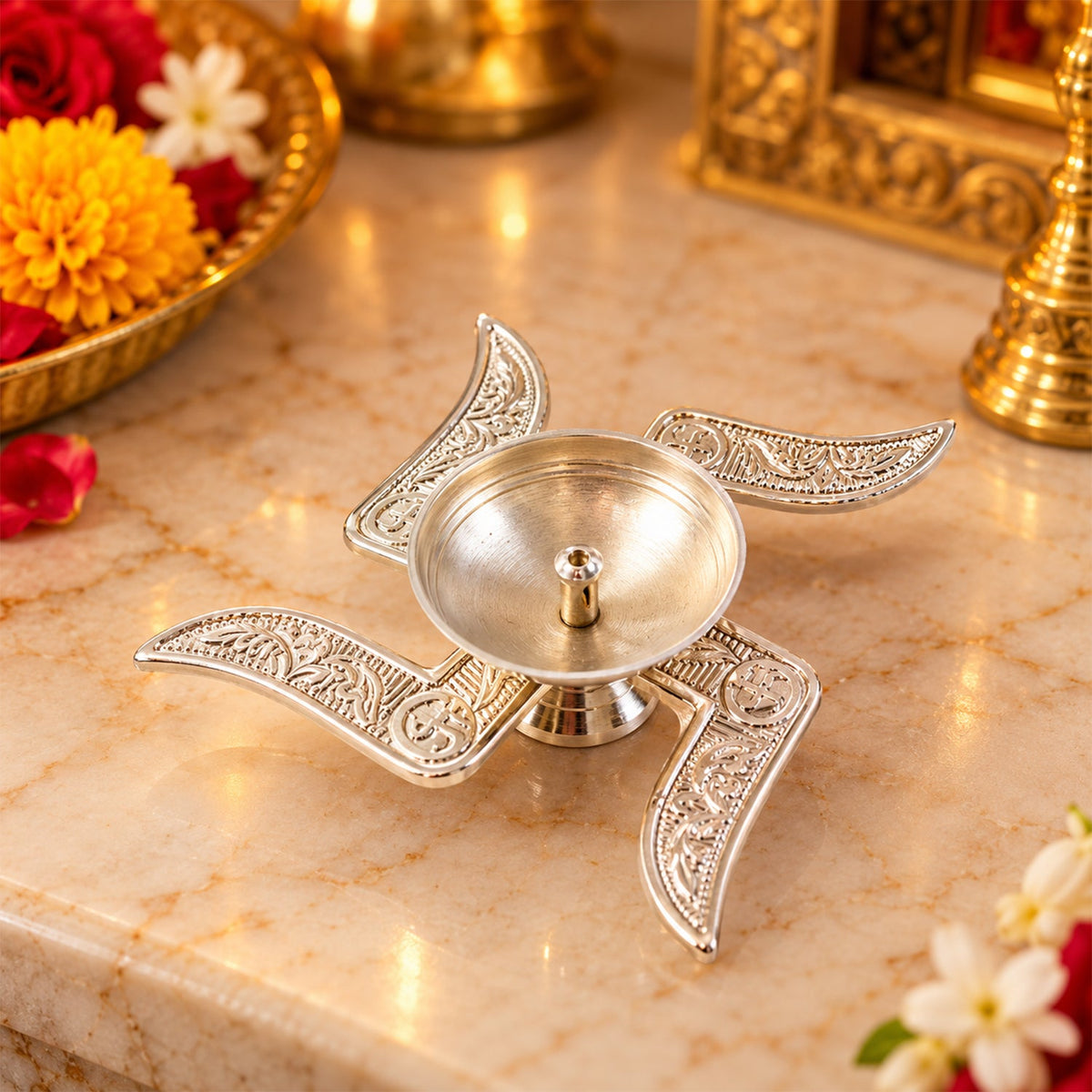A silver Swastik diya with intricate patterns rests on a marble surface with flowers and a golden tray in the background.