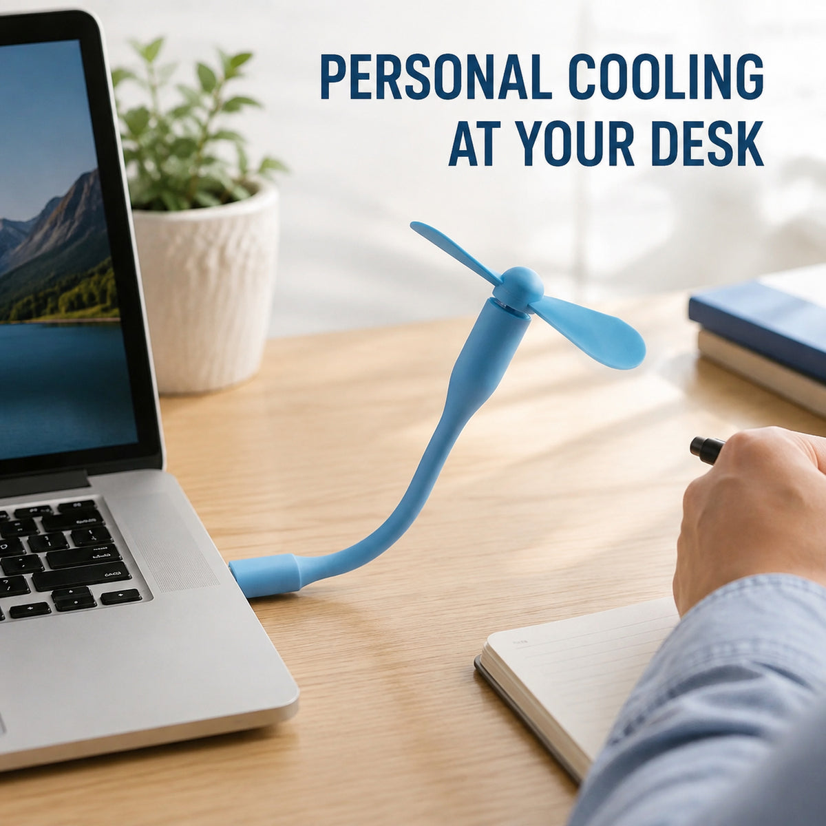 A blue flexible USB fan is plugged into a laptop on a wooden desk with a person's hand writing in a notebook.