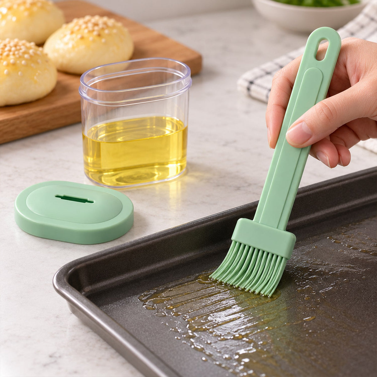 A hand uses a light green silicone basting brush to spread oil on a dark baking sheet, with a clear oil container and baked