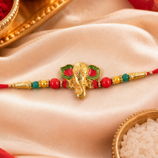A gold Ganesha head charm with red and green enamel accents sits on a red thread, flanked by red, green, and gold beads, on