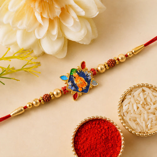 A red thread with gold beads and a blue square charm featuring an orange Hanuman sits on a beige surface with flowers, rice,