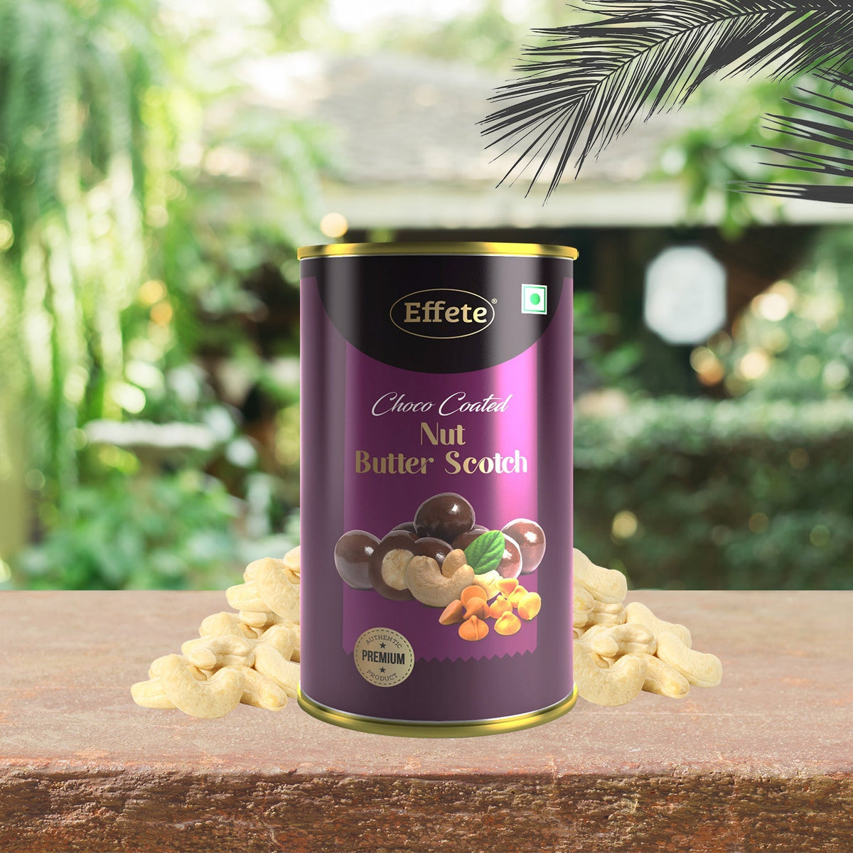A purple and gold tin of chocolate-coated nuts sits on a wooden surface, surrounded by whole cashews and palm leaves.