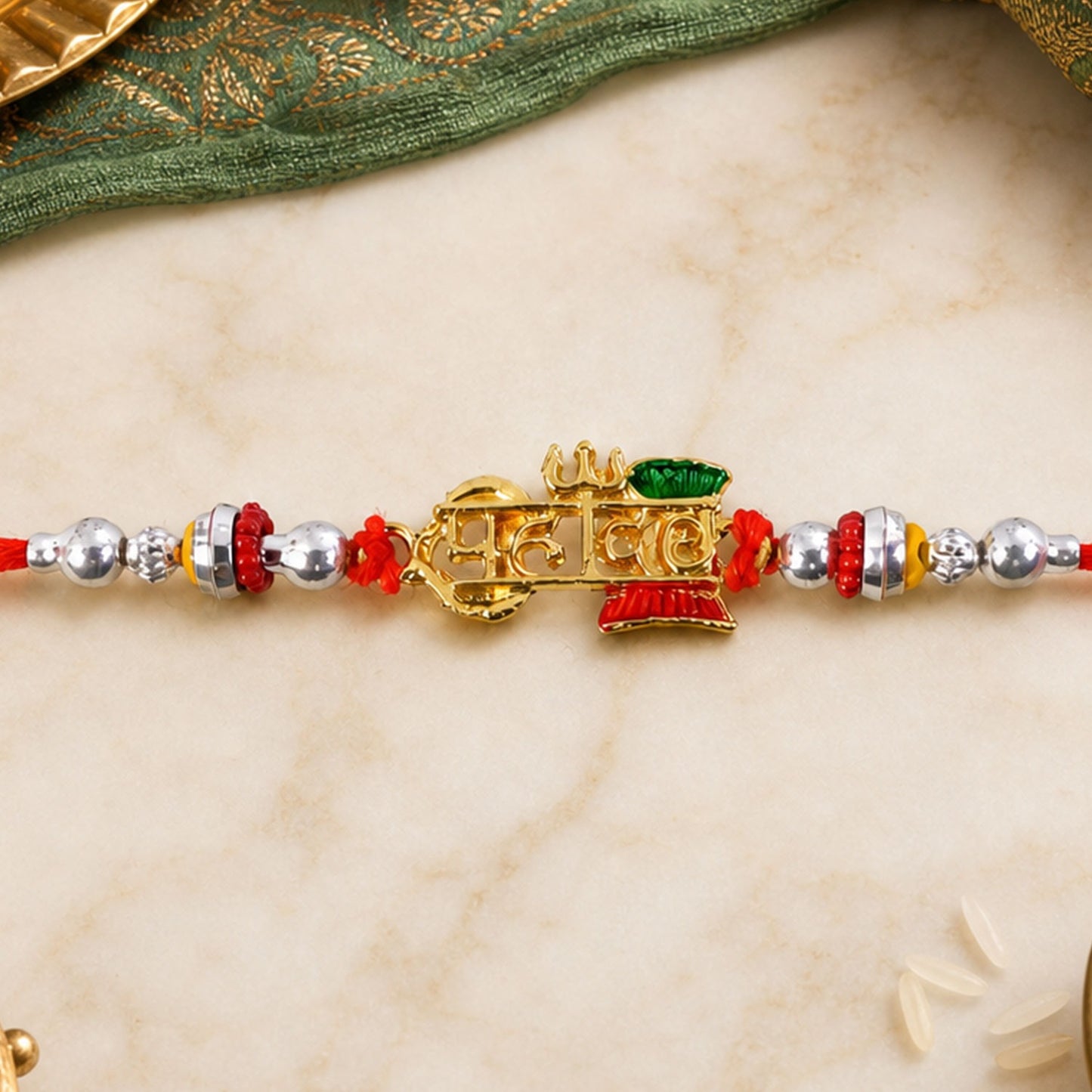 A red thread with silver beads and a gold-colored centerpiece with green and red accents rests on a light marble surface.