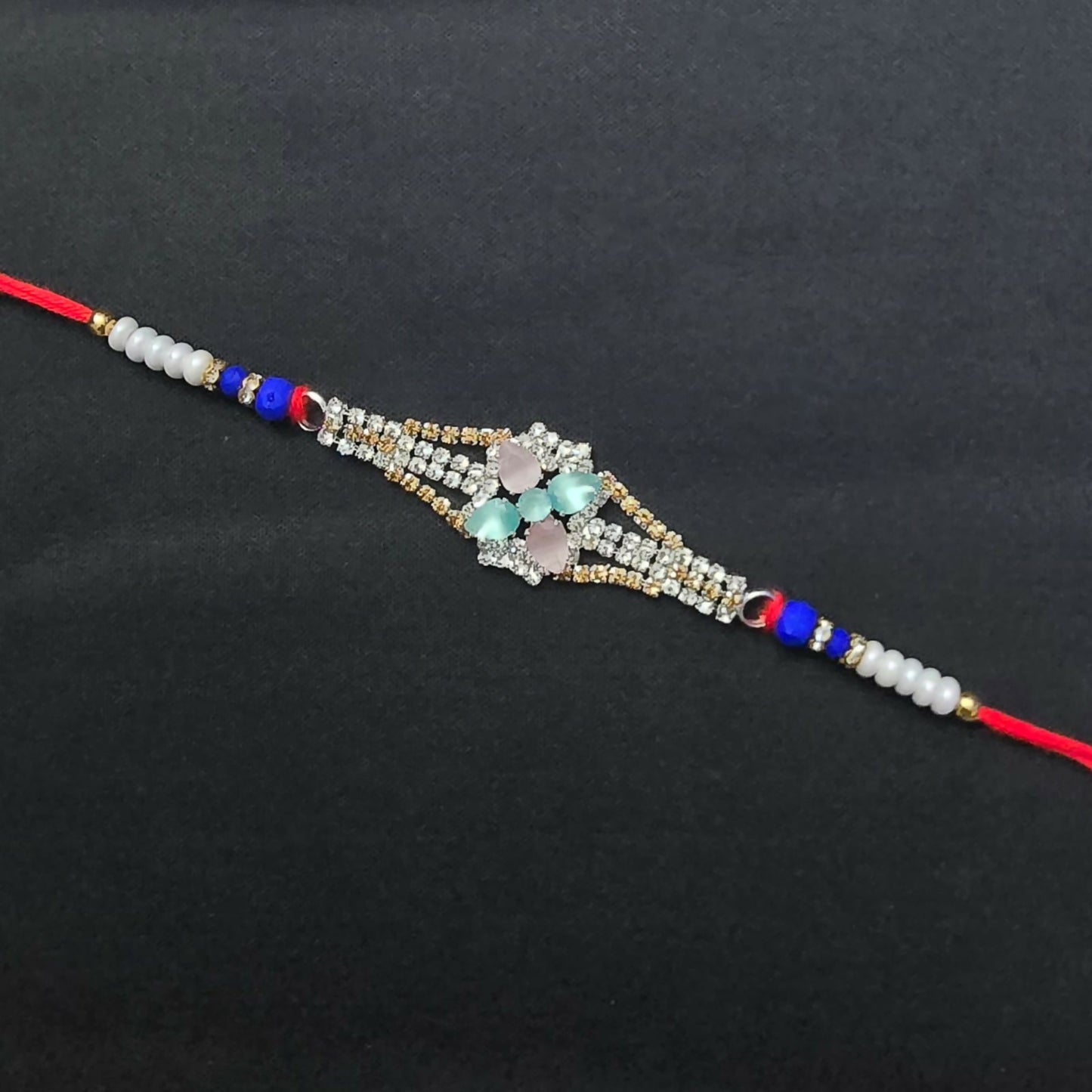 A red thread bracelet with a central diamond-studded floral design featuring pink and blue-green crystals, on a plain black