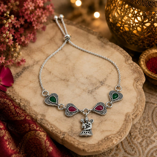 An oxidized silver bolo bracelet with ruby and emerald teardrop stones and a charm of two figures is displayed on a wooden