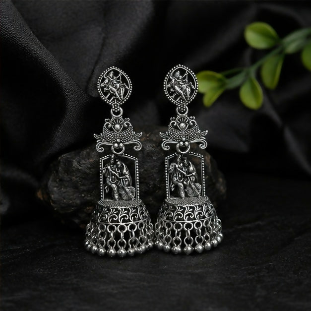A pair of oxidized silver jhumka earrings with Radha Krishna temple design on a black fabric background.