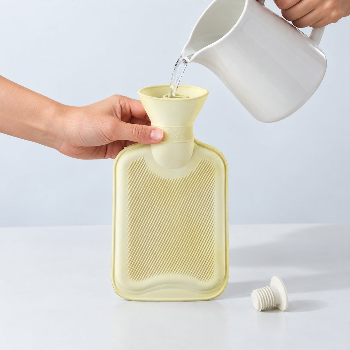 Hands pour water from a white pitcher into a light yellow hot water bottle, with its white stopper nearby.