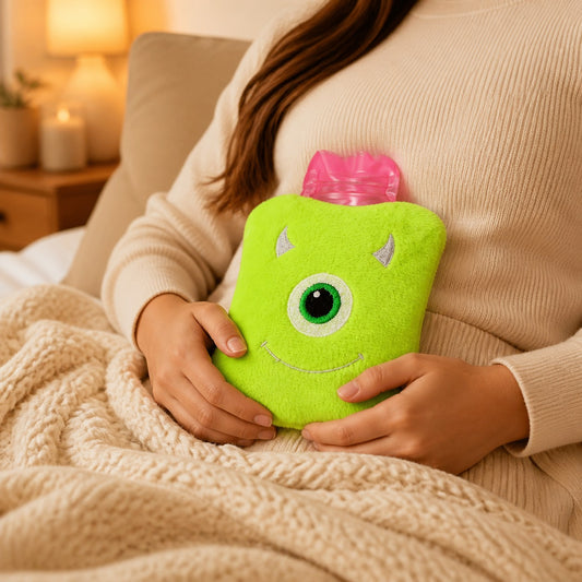 A person in a cream sweater holds a green monster hot water bag on their stomach while sitting on a bed.
