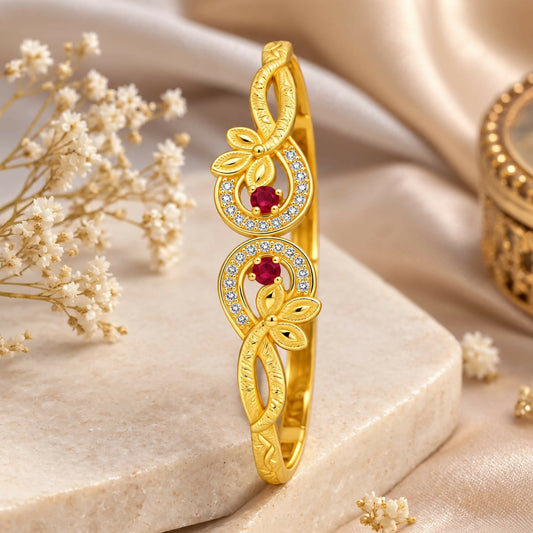A gold-plated bracelet with two floral swirls, each featuring a ruby surrounded by small diamonds, rests on a white surface.