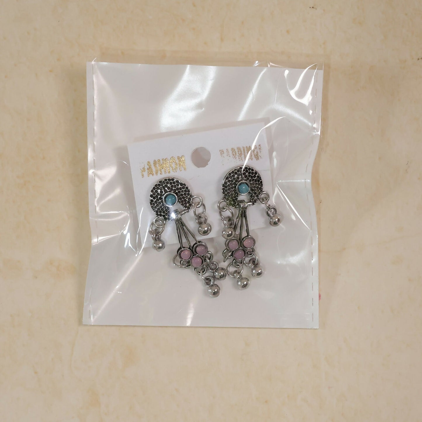 The oxidised silver long drop earrings with pink and turquoise stones and ghungroo bells are packaged in a clear plastic bag.