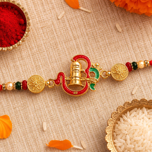 A red and gold enamel Shivling Rakhi with beads and gold coin accents lies on a beige fabric with scattered rice and flower