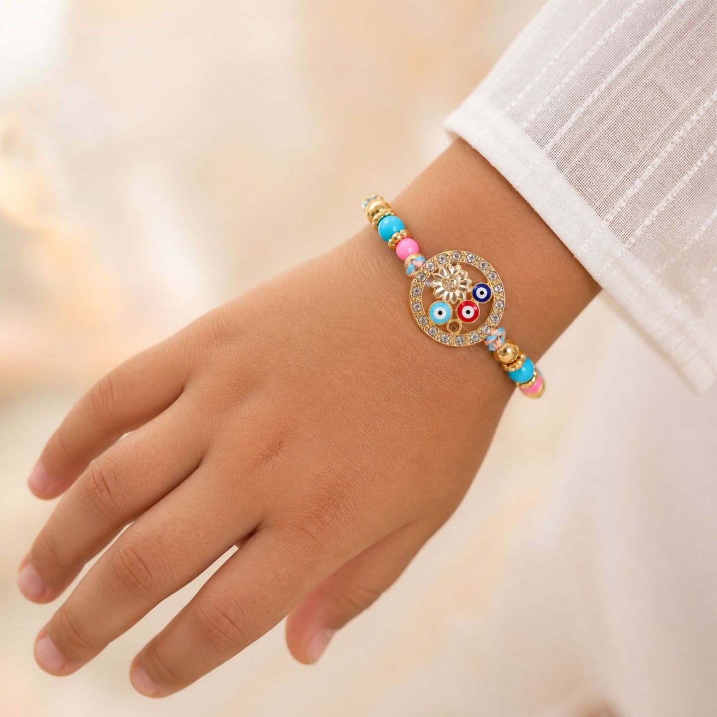 A child's hand wearing a colorful Rakhi bracelet with evil eye beads and a gold flower charm.
