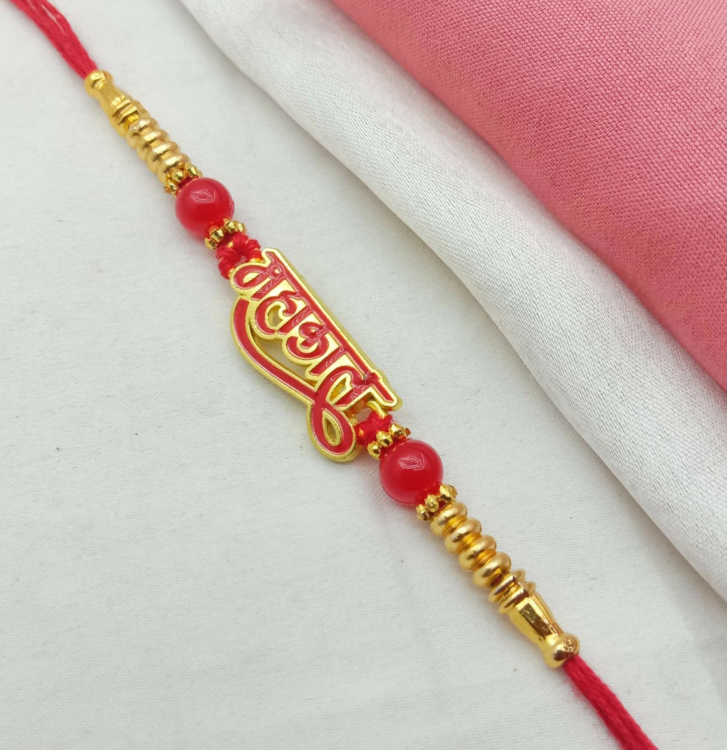 A red thread with a gold and red Mahakal calligraphy charm, red beads, and gold accents lies on a white and pink fabric