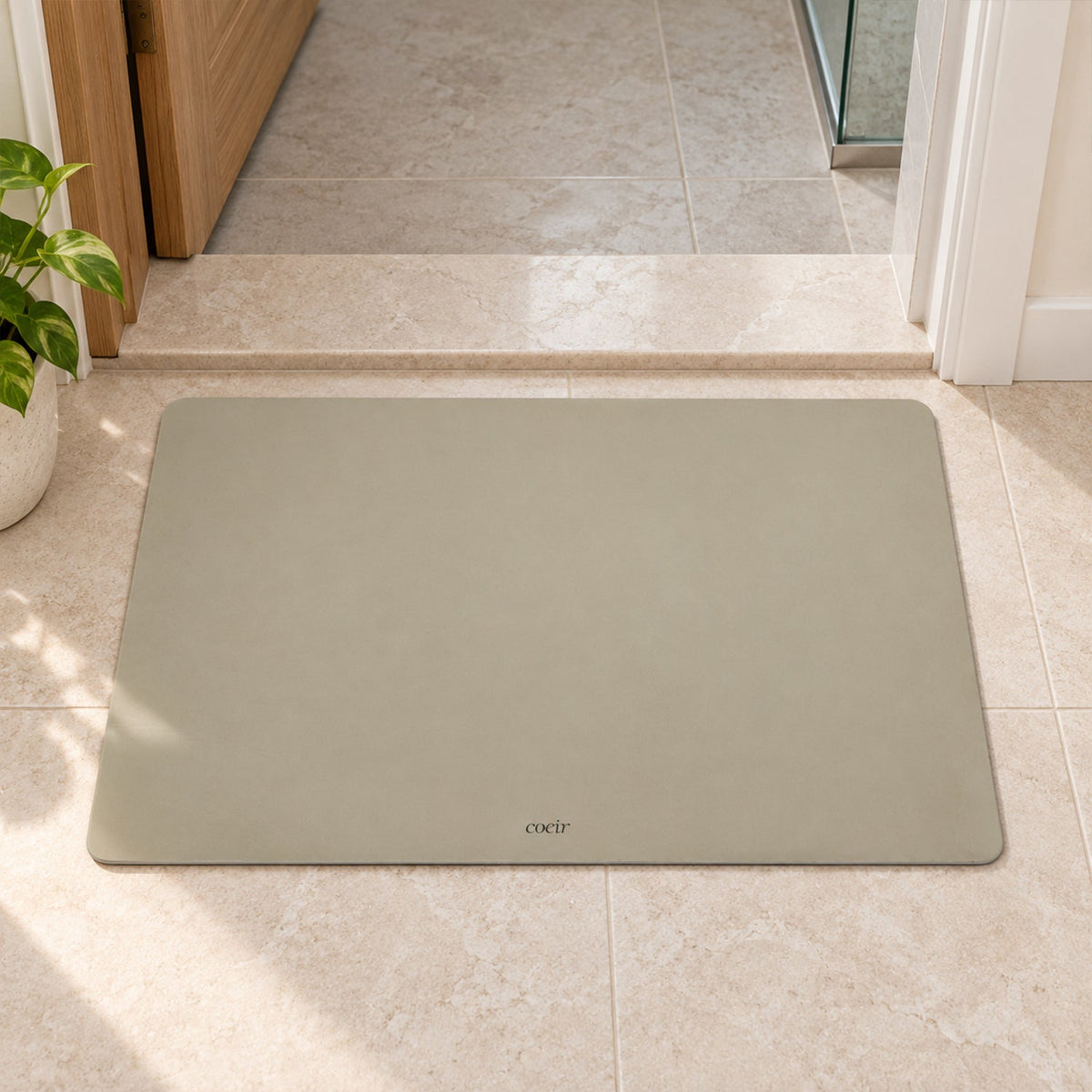 A light beige rectangular floor mat rests on tiled flooring near a doorway with a plant.