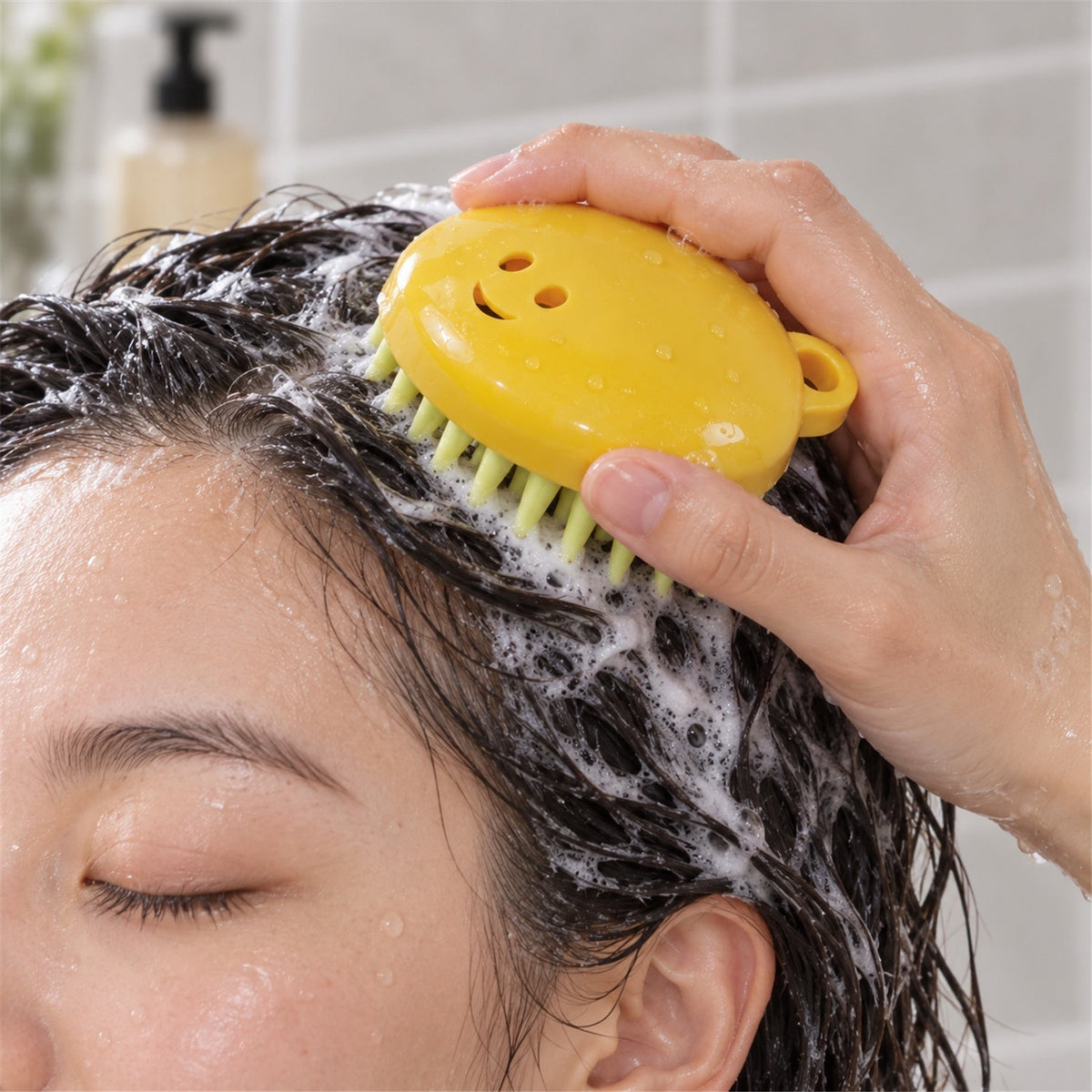 A person's hand holds a yellow silicone brush with light green bristles, massaging shampoo into dark hair.