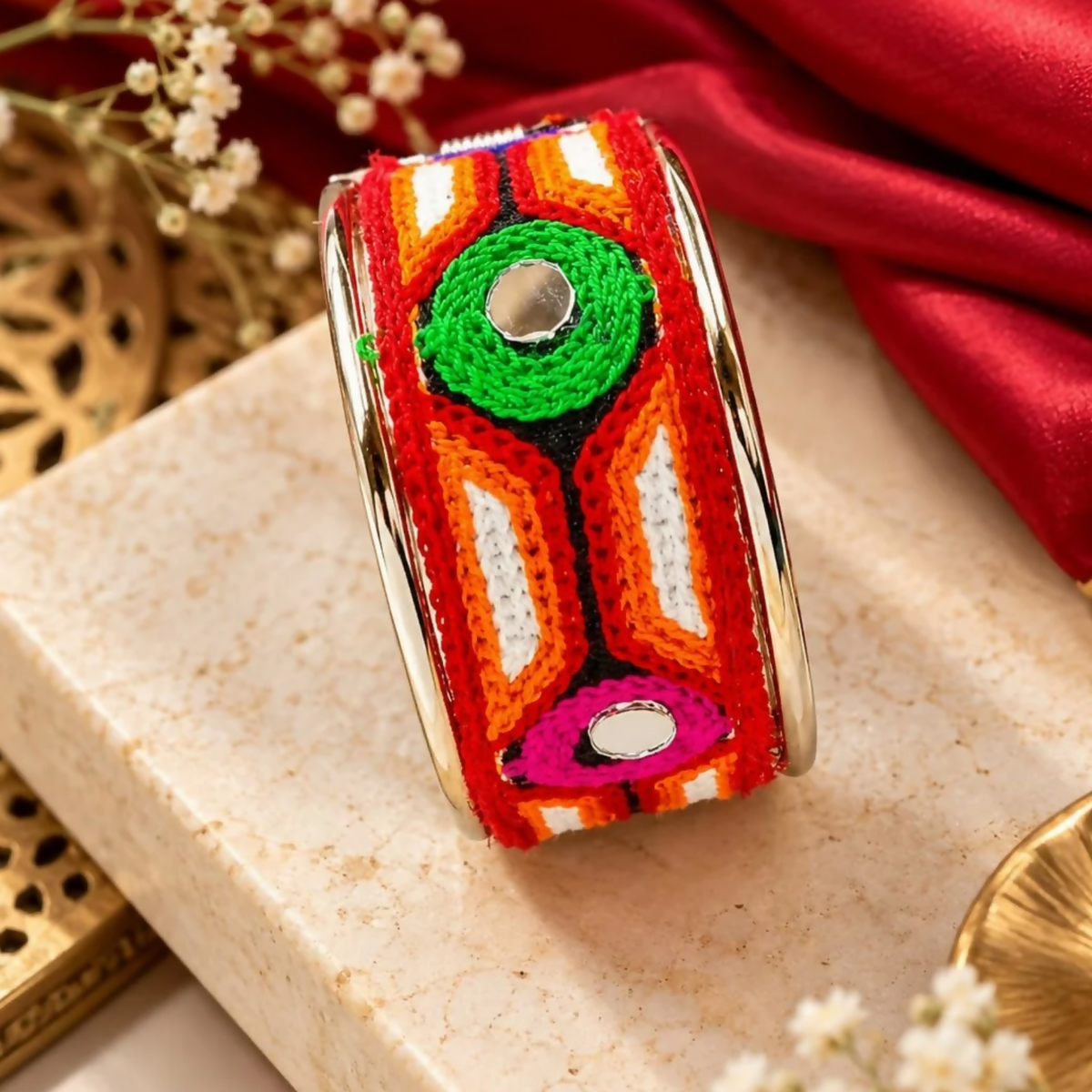A vibrant red, orange, white, and green embroidered cuff bracelet with two small mirrors, resting on a light brown surface.
