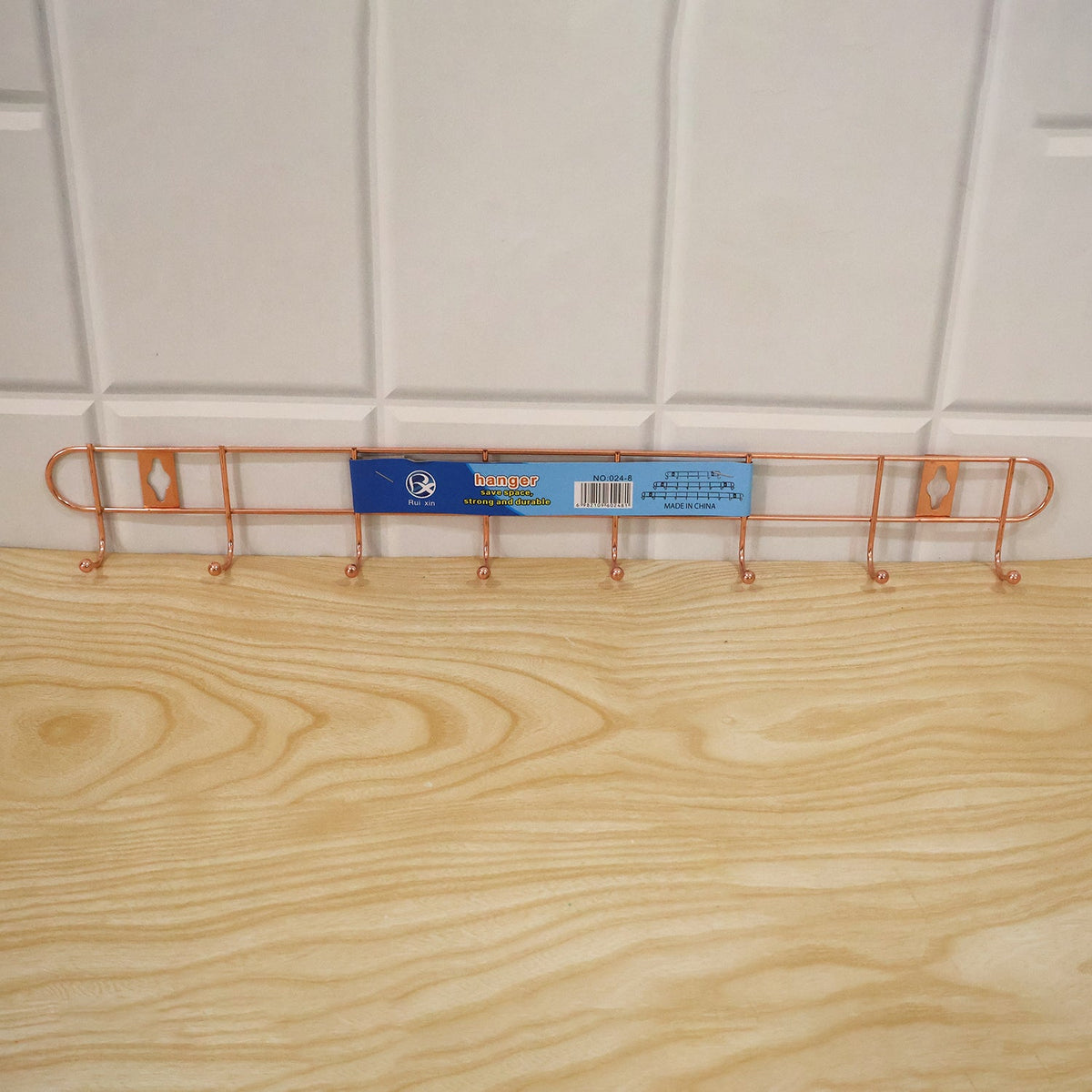 A rose gold metal wall hanger with eight hooks has a product label attached to it, resting on a light wood surface.