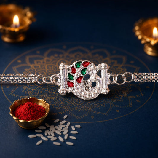 A silver chain Rakhi with a peacock design, featuring red and green enamel, rests on a blue patterned surface with two lit
