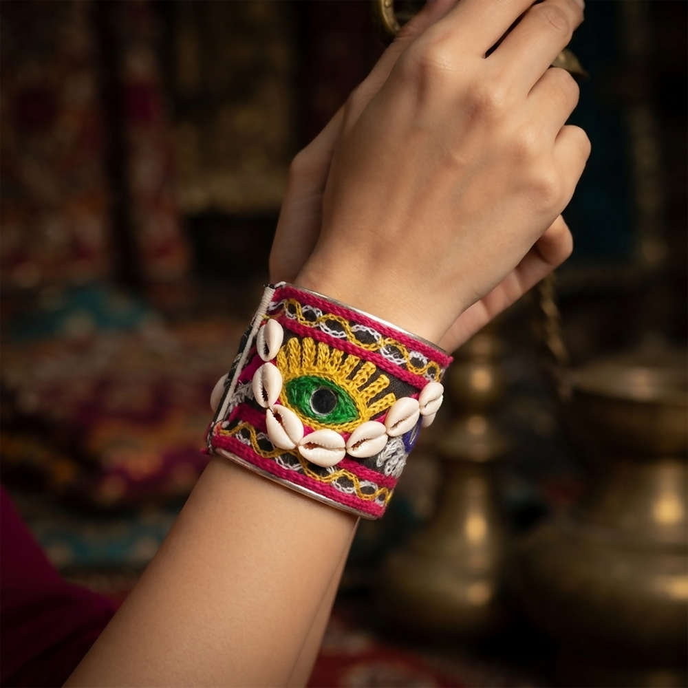 A person's arm wearing a wide silver bangle with black fabric, colorful embroidery, and cowrie shells.
