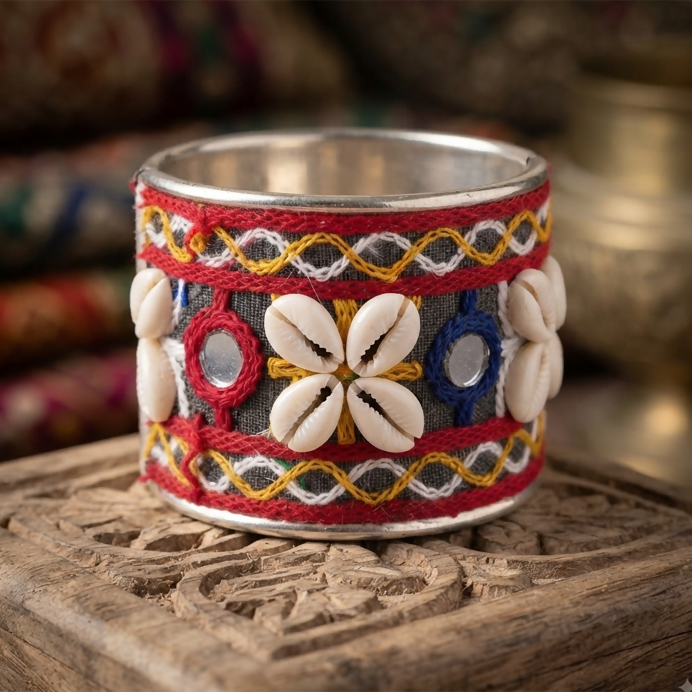 A wide silver cuff bracelet with colorful embroidered patterns, small round mirrors, and white cowrie shells sits on a