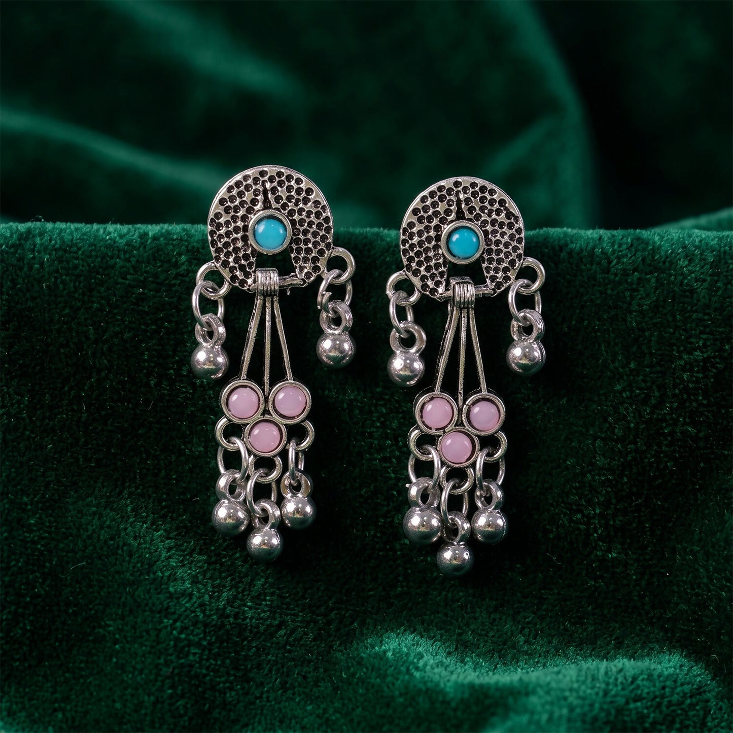 The oxidised silver long drop earrings with pink and turquoise stones and ghungroo bells are displayed on a dark green
