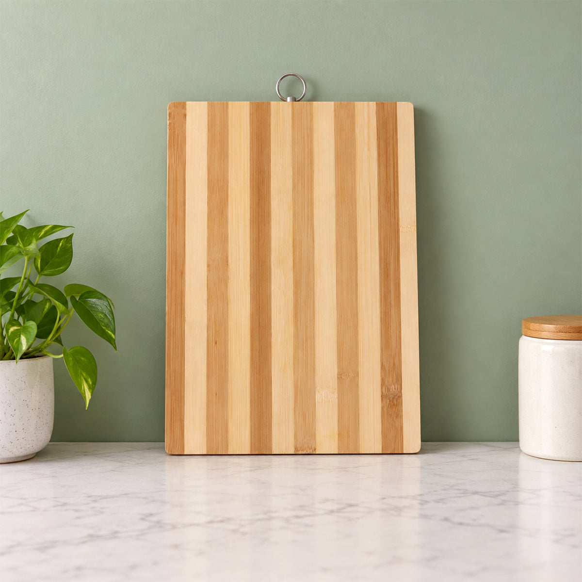 A rectangular bamboo cutting board with light and dark stripes hangs on a green wall above a marble counter.