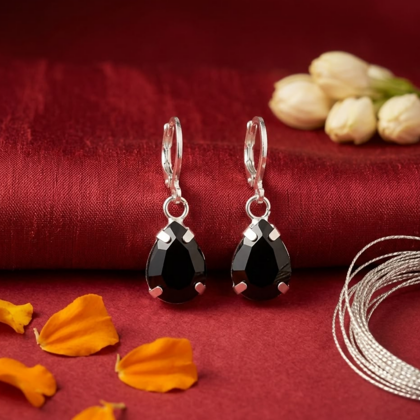 Two black teardrop crystal earrings on a red fabric with orange petals and silver string, and white flowers in the