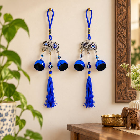 Two blue elephant evil eye wind chimes with bells and tassels hanging on a light wall next to a plant and wooden decor.