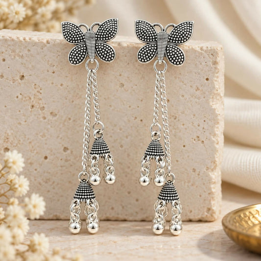 A pair of silver butterfly earrings with long chains and small jhumka bells, resting on a light-colored stone block.