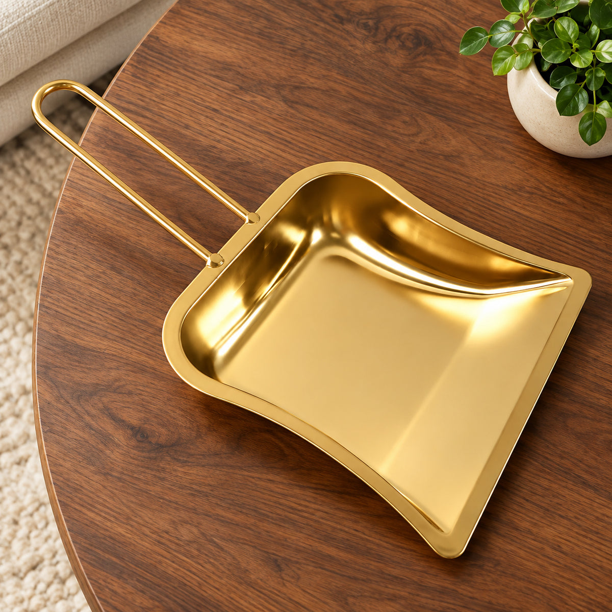 A gold-tone metal dustpan with a loop handle sits on a dark wooden coffee table beside a small green plant.