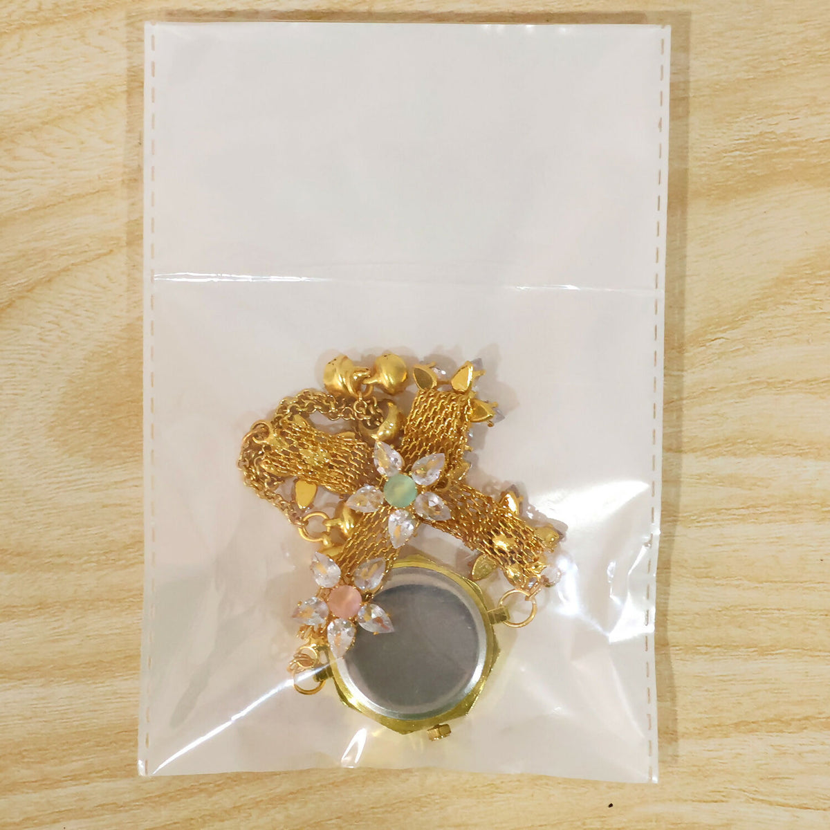 The gold-tone bracelet watch, folded, is sealed inside a clear plastic bag on a light wooden surface.