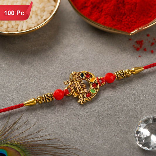 A red thread bracelet with a golden Lord Krishna charm, red beads, and golden accents rests on a grey surface with rice and